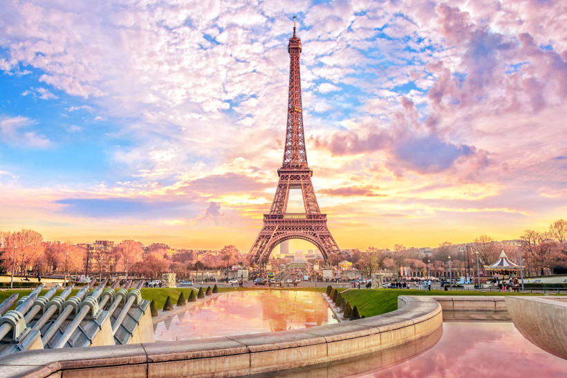 Frankreich Der Eiffelturm in Paris bei Sonnenuntergang, umgeben von einem farbenprächtigen Himmel in Rosa, Orange und Blau. Im Vordergrund spiegeln sich die Farben im Wasser eines flachen Beckens. Links sind Wasserkanonen der Fontänenanlage zu sehen, rechts ein klei