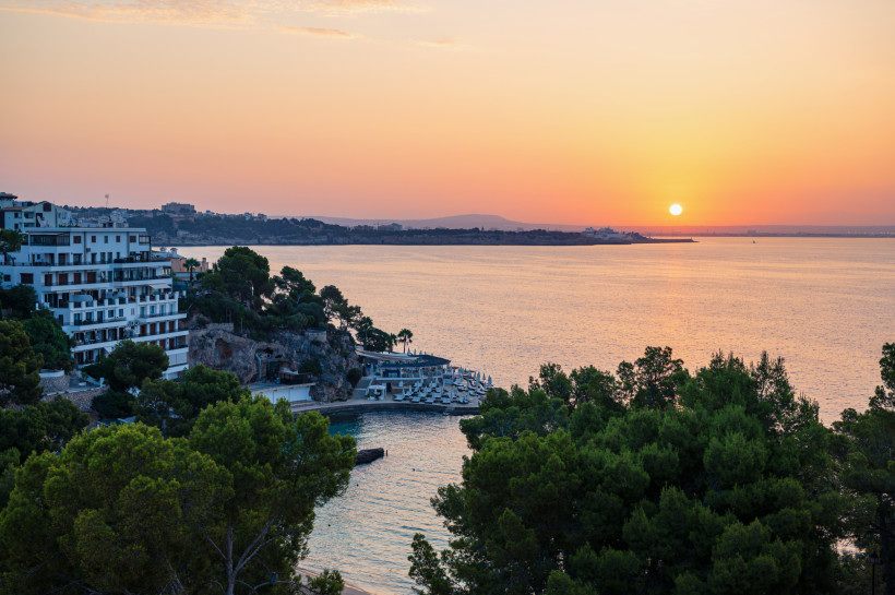 Sonnenuntergang über der Bucht von Illetas mit Hotelgebäude am Fels, Strandliegen und Pinien im Vordergrund
