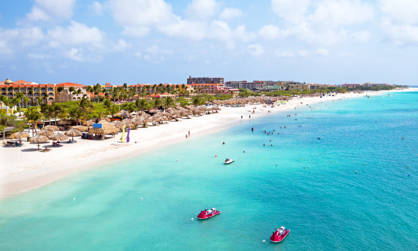 Panoramablick auf den weißen Sandstrand von Eagle Beach auf Aruba. Im türkisblauen Wasser sind Jet-Skis unterwegs, während zahlreiche Menschen im Wasser baden oder am Ufer spazieren. Am Strand stehen Palapa-Sonnenschirme und Liegen. Dahinter reihen sich R
