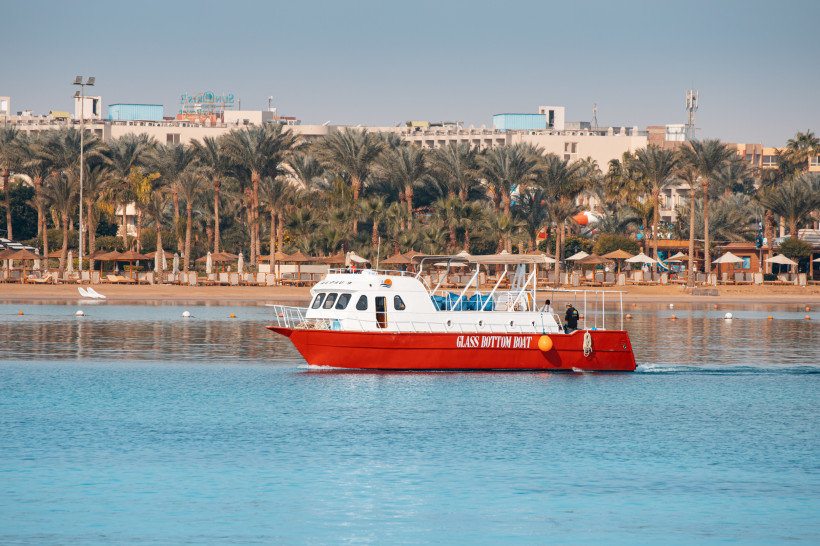 Ein rotes Glasbodenboot fährt ruhig entlang der Küste von Hurghada, im Hintergrund Palmen und Hotelanlagen.