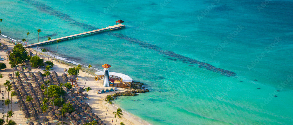 Dominikanische Republik Strand mit Palmen, kleinen Strohhütten und einem blau-weiß gestreiften Leuchtturm mit Steg im türkisfarbenen Meer