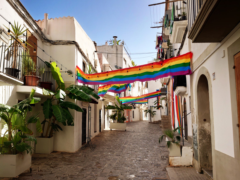 Ibiza Eine schmale Gasse in Ibiza-Stadt, gesäumt von weiß gestrichenen Häusern mit Balkonen und Pflanzen. Mehrere große Regenbogenflaggen hängen quer über die Straße, als festlicher Schmuck. Die Szene ist sonnig und friedlich.