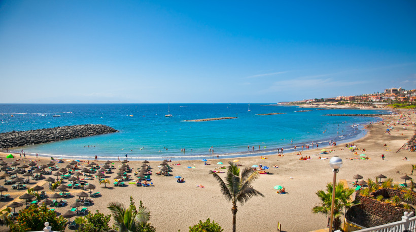 Badeparadies Playa de las Américas auf Teneriffa Gut besuchter Sandstrand in Playa de las Américas auf Teneriffa mit türkisblauem Meer, Sonnenschirmen, Badenden und Palmen unter blauem Himmel.