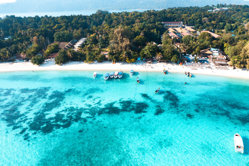 Thailand Pattaya Luftaufnahme eines tropischen Resorts mit weißen Sandstränden und türkisblauem Wasser. Mehrere Boote ankern vor der Küste, dahinter dichte grüne Vegetation und Hotelanlagen.