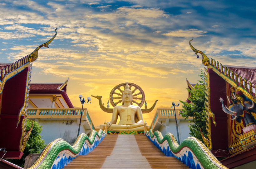Thailand - Koh Samui  Das Bild zeigt eine monumentale goldene Buddha-Statue, die auf einer Plattform über einer langen, breiten Treppe thront. Die Treppe ist bunt gestaltet, mit wellenartigen grünen und blauen Elementen am Rand, die an Nagas – mythische Schlangenwesen – erinn
