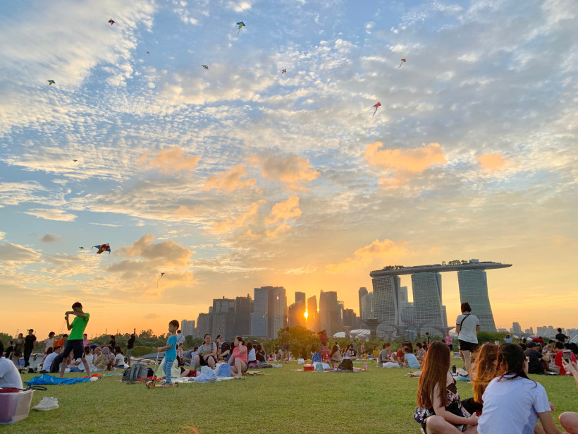 Singapur Menschen genießen ein Picknick auf einer großen Wiese im Marina Barrage Park in Singapur. Die Skyline mit dem markanten Marina Bay Sands Hotel ist im Hintergrund sichtbar. Der Himmel ist bunt gefärbt durch die untergehende Sonne, am Himmel fliegen mehrere