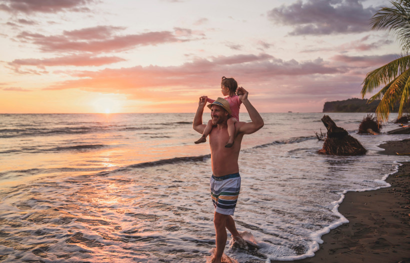 Costa Rica, Familienurlaub  Vater trägt seine Tochter auf den Schultern beim Spaziergang am Strand während eines farbenfrohen Sonnenuntergangs