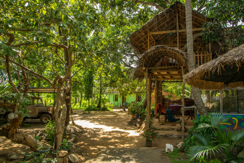 Naturnahe Unterkunft in Sri Lanka mit einem rustikalen Baumhaus aus Holz und Palmendach, eingebettet in tropisches Grün. Im Vordergrund Bäume, Pflanzen und ein unbefestigter Weg. Links ein Jeep unter einem Unterstand, rechts Sitzgelegenheiten unter dem Ba