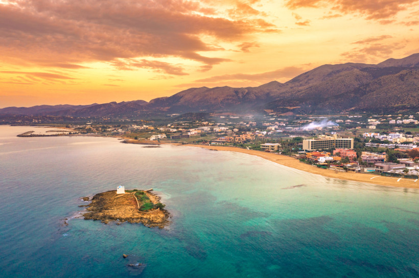 Küstenlandschaft bei Malia mit Sandstrand, kleinem Felseninselchen und Bergen im Hintergrund bei Sonnenuntergang