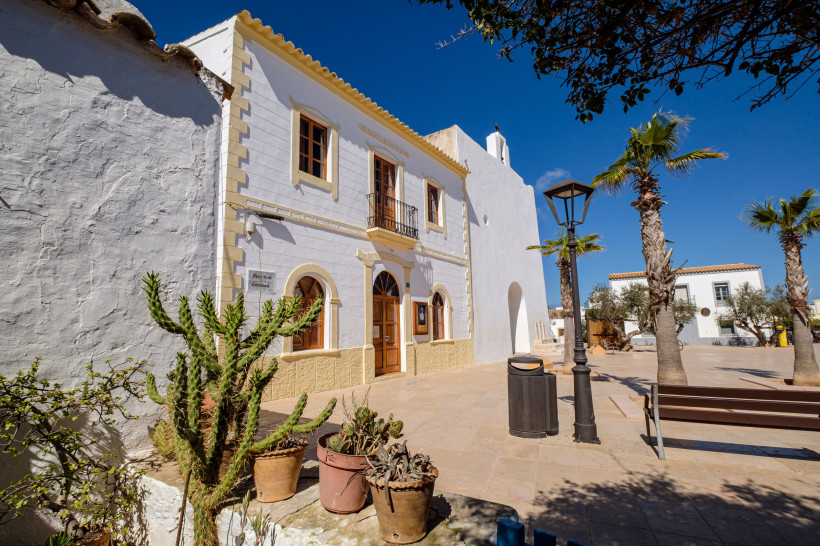 Formentera Helle, historische Gebäude ( Städtchen Sant Francesc de Formentera ) mit weißer Fassade und braunen Fensterläden in einem sonnigen Dorfplatz auf Formentera. Im Vordergrund stehen Blumentöpfe mit Kakteen, eine Holzbank und Palmen säumen den Platz. Die Szen