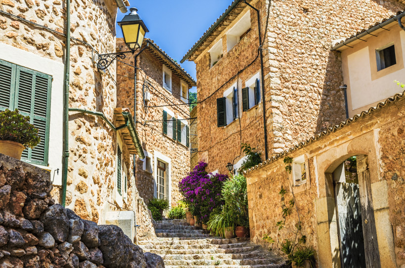 Mallorca - Fornalutx Eine idyllische enge Gasse in einem mallorquinischen Dorf mit traditionellen Sandsteinfassaden und grünen Fensterläden. Bunte Blumentöpfe mit Bougainvillea und anderen mediterranen Pflanzen schmücken die Treppenstufen. Im Hintergrund ein wolkenloser, blau
