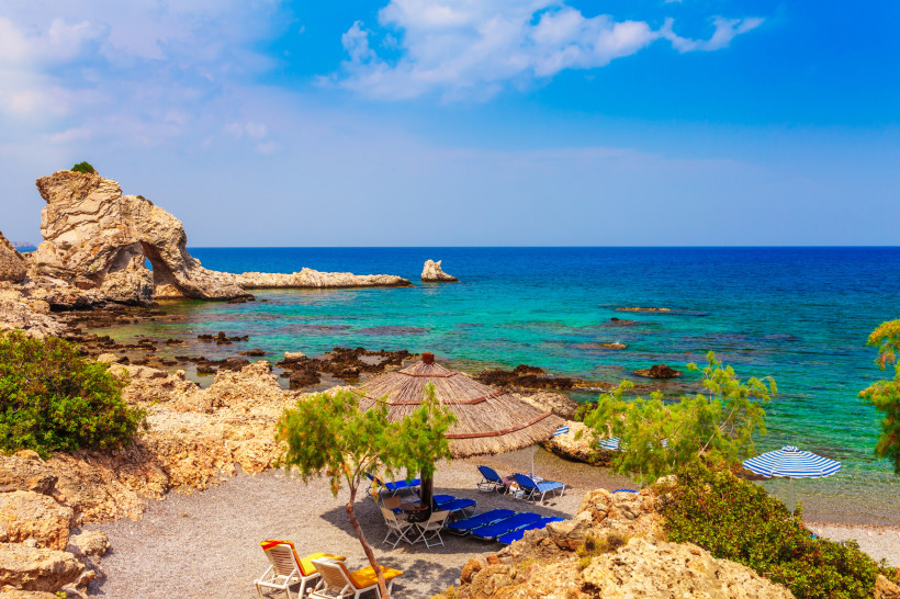 Kleine felsige Badebucht auf Rhodos mit Strohdach-Sonnenschirmen, Liegen und kristallklarem türkisblauem Meer vor zerklüfteter Küste